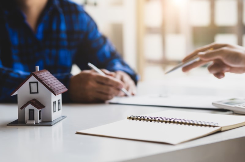 Acompanhe o processo de análise e aprovação | Miniatura de casa sobre a mesa enquanto duas pessoas preenchem documentos | Minha Casa Minha Vida | Blog da Tenda 