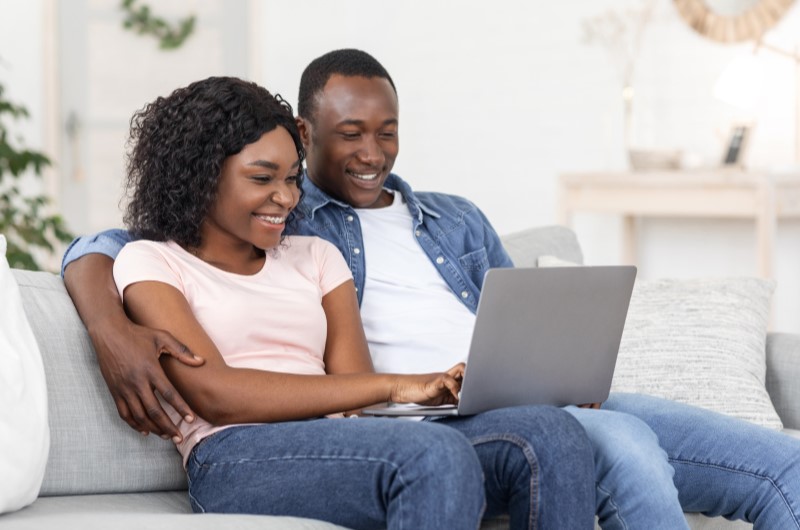 Ipanema | Casal sorridente usando o notebook no sofá da sala de estar | Trilha da Conquista | Blog da Tenda