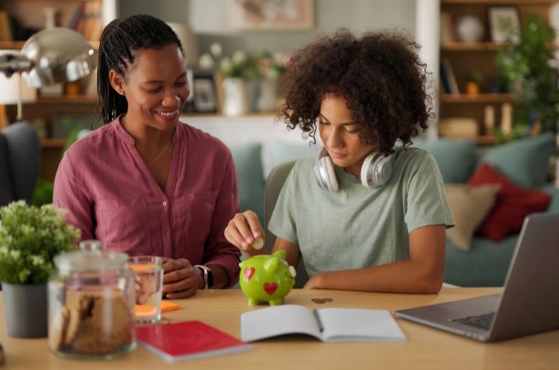 O que é educação financeira? | Mãe ajudando filho a colocar moeda em um cofrinho verde, em um ambiente aconchegante | Eu Dou Conta | Blog da Tenda 