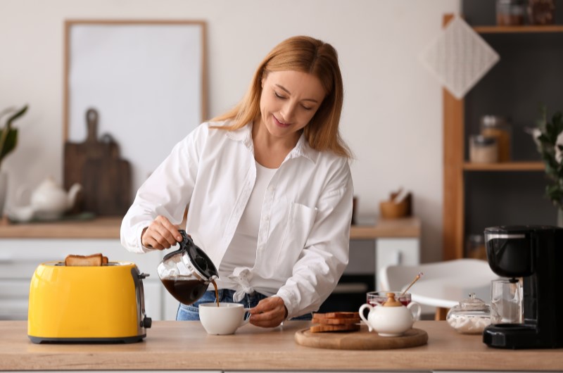 Acessórios | Mulher servindo café em uma xícara ao lado de uma torradeira amarela | Casa e Decoração | Blog da Tenda 