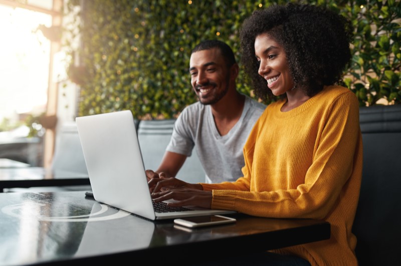 Como mobiliar um apartamento sem gastar muito | Casal sentado em cafeteria, sorrindo enquanto utiliza um notebook para trabalhar ou pesquisar | Casa e Decoração | Blog da Tenda 