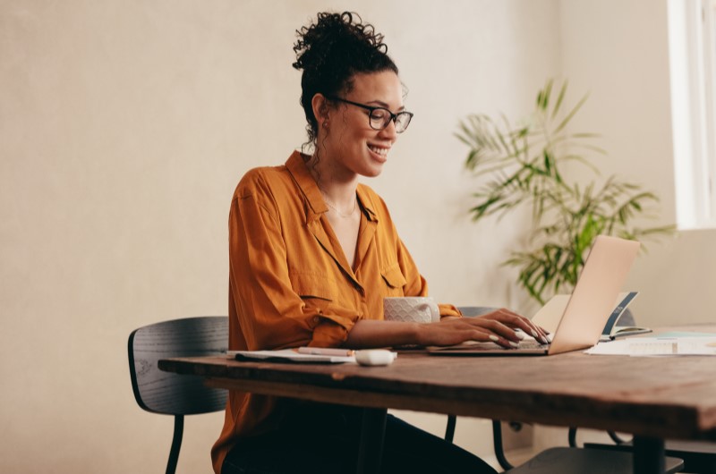 Como se cadastrar no Minha Casa Minha Vida | Mulher sorridente trabalhando em um laptop em sua mesa | Minha Casa Minha Vida | Blog da Tenda 