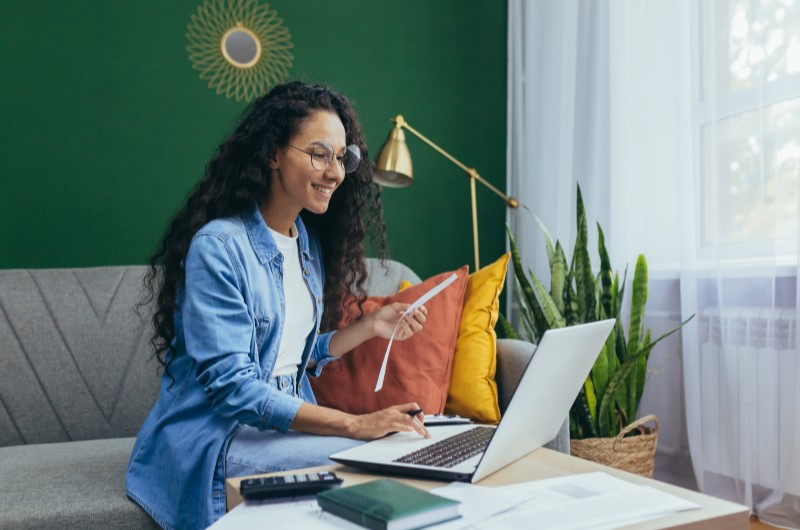 Prepare-se para um novo estilo de vida | Mulher sorrindo enquanto segura um papel e trabalha em um notebok na sala | Trilha da Conquista | Blog da Tenda 