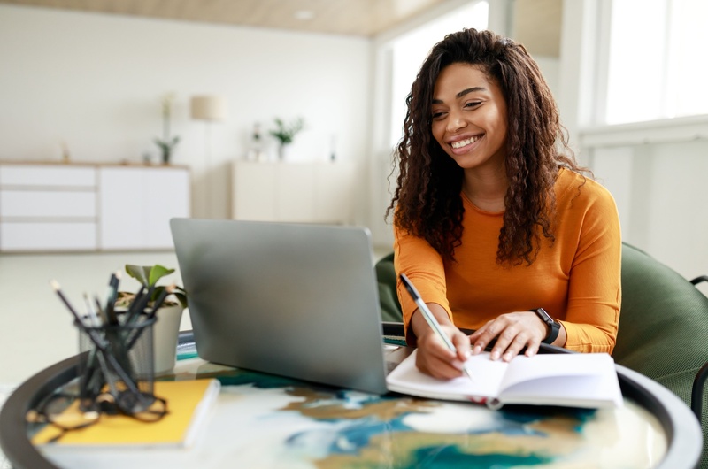 Como é morar em condomínio residencial | Jovem sorridente escrevendo em um caderno com um notebook ao lado | Viver em Condomínio | Blog da Tenda 