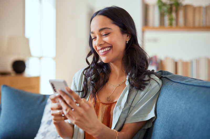 Acompanhe o seu progresso | Mulher sorridente usando celular no sofá | Eu Dou Conta | Blog da Tenda 