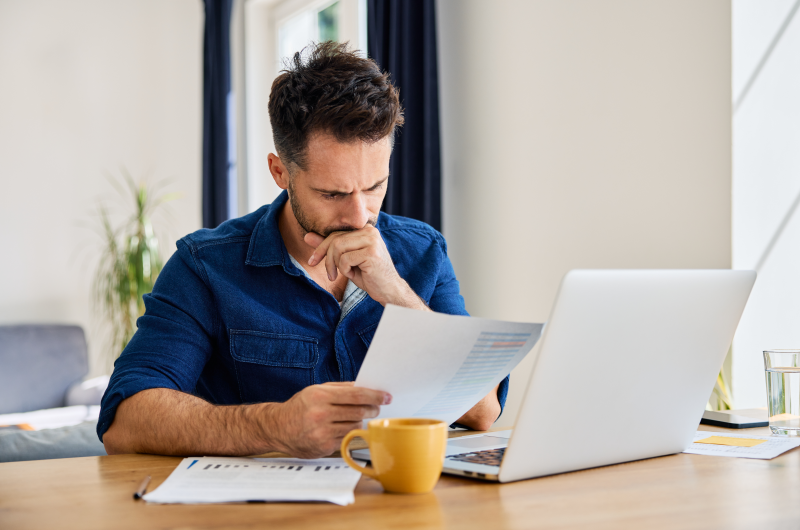 O que é um condômino inadimplente | Homem concentrado revisando documentos ao lado de um notebook e uma xícara de café | Viver em Condomínio | Blog da Tenda 