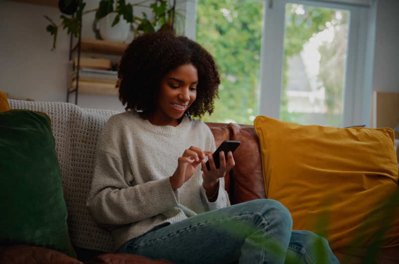 Dúvidas comuns sobre a Compra Planejada | Mulher sorrindo enquanto usa o celular, sentada em um sofá confortável em casa | Eu Dou Conta | Blog da Tenda 