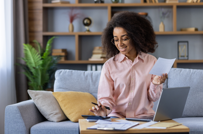 Faça um planejamento financeiro para juntar dinheiro | Mulher revisando documentos financeiros no sofá, ao lado de um notebook | Eu Dou Conta | Blog da Tenda 