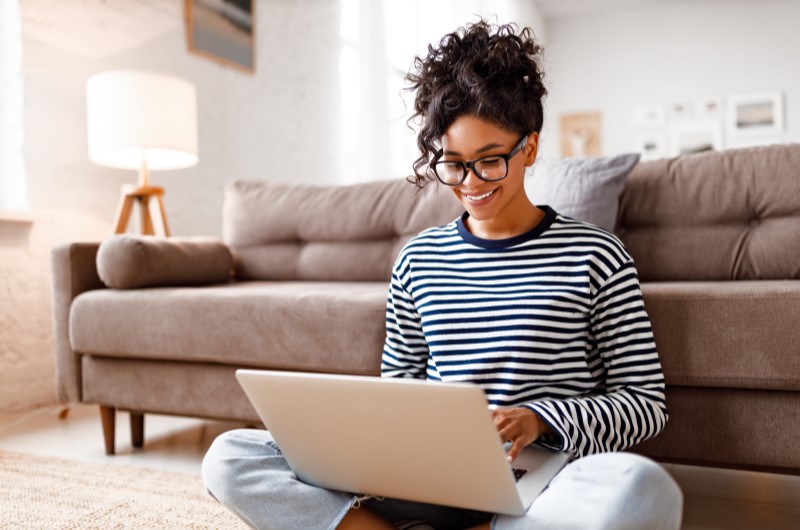 Vantagens do financiamento para servidor público | Mulher sorridente usando notebook sentada no chão de casa | Trilha da Conquista | Blog da Tenda