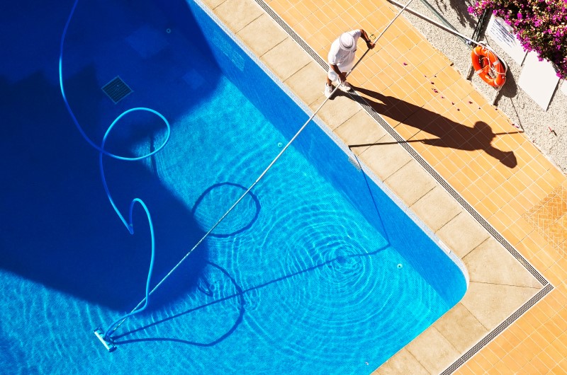 Entenda as regras de segurança e higiene | Pessoa limpando uma piscina com cabo longo e mangueira azul | Viver em Condomínio | Blog da Tenda 