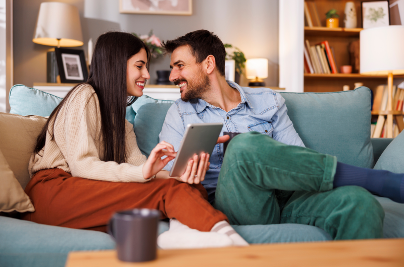 Onde fica o bairro Jaçanã | Casal sorridente usando um tablet no sofá da sala | Onde Morar | Blog da Tenda 