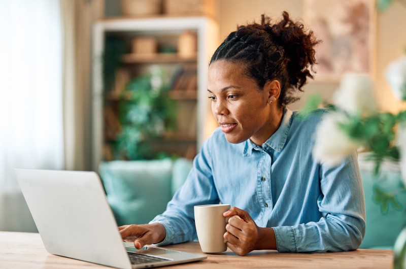 Score de crédito baixo | Mulher sorrindo enquanto usa um notebook e segura uma xícara | Mulher sorrindo enquanto usa um notebook e segura uma xícara | Trilha da Conquista | Blog da Tenda 