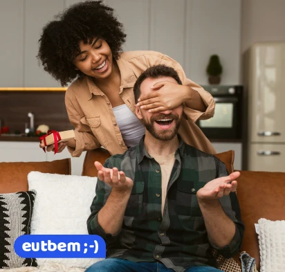 Mulher sorrindo enquanto cobre os olhos do parceiro com um presente nas mãos, representando momentos de felicidade e conquistas alcançadas com o Consórcio Eutbem e a Compra Planejada.