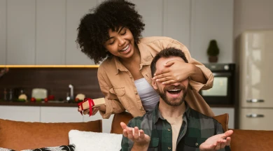 Mulher sorrindo enquanto cobre os olhos do parceiro com um presente nas mãos, representando momentos de felicidade e conquistas alcançadas com o Consórcio Eutbem e a Compra Planejada.