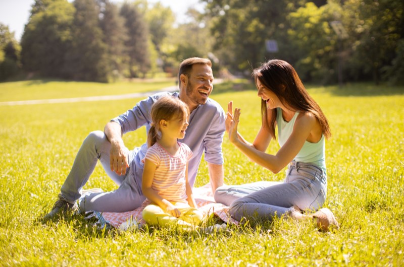 Passeios ao ar livre | Foto de uma família sorridente sentada na grama de um parque | Economia e renda extra | Eu Dou Conta 