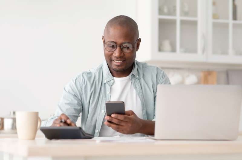 O que é independência financeira? | Foto de um homem sorridente olhando para o celular e usando a calculadora | Economia e renda extra | Eu Dou Conta 