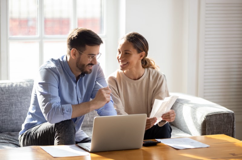 Quais são as vantagens? | Foto de um casal sorridente conversando e usando o notebook | Dúvidas sobre dívidas | Eu Dou Conta 