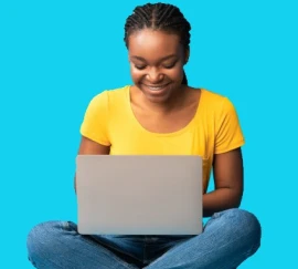 Mulher sorridente usando uma camiseta amarela, sentada com as pernas cruzadas enquanto utiliza um laptop, com fundo azul vibrante.