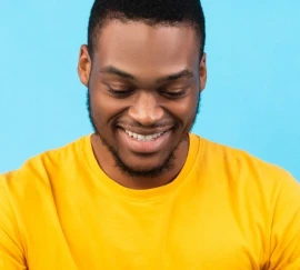 Homem sorrindo usando uma camiseta amarela, com fundo azul claro, olhando para baixo com expressão de satisfação.